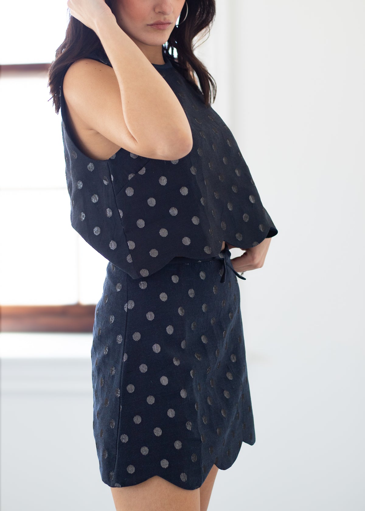 A woman wearing a sleeveless dark navy dress with a silver polka dot pattern, standing in a brightly lit room.
