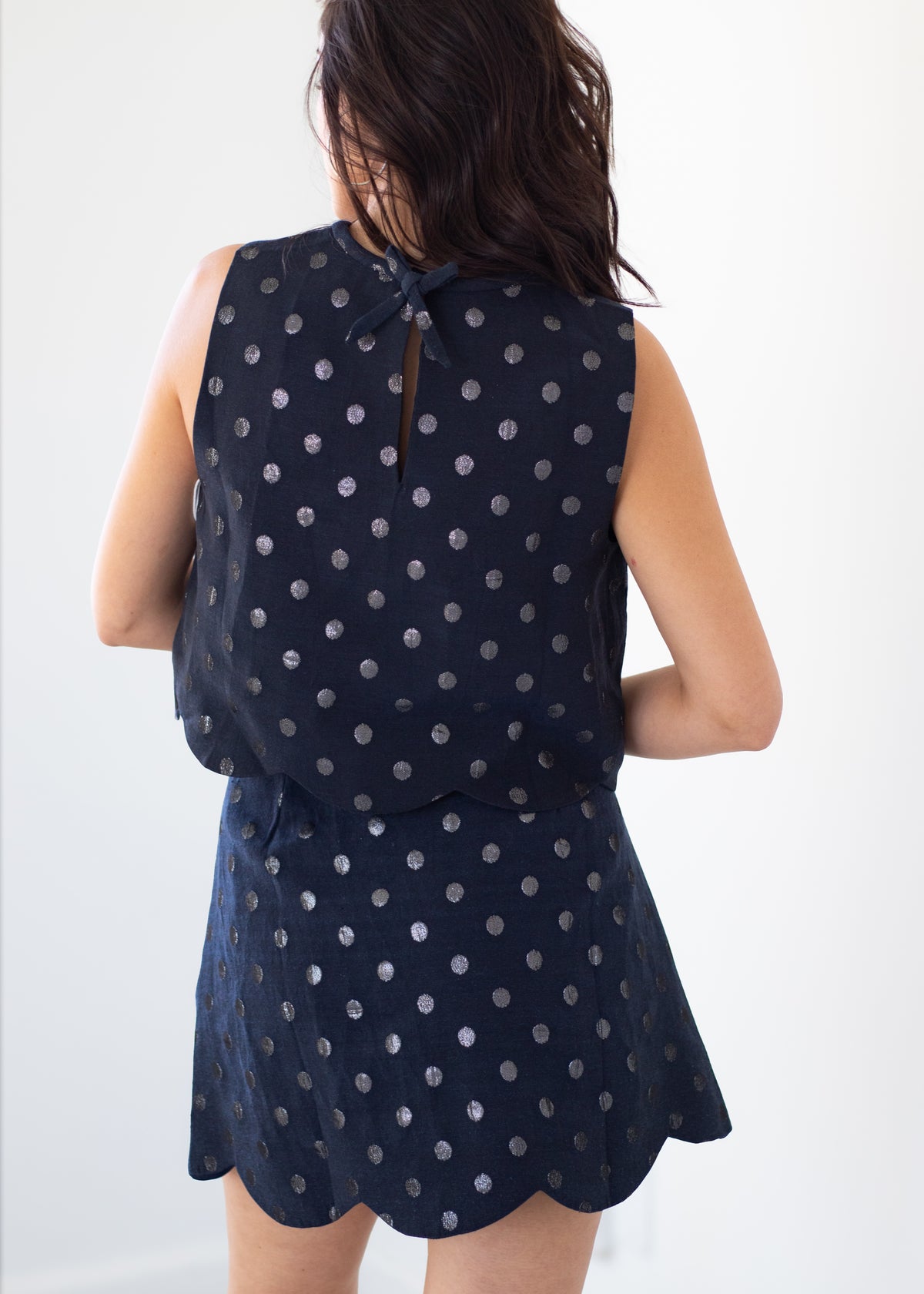 A woman shown from the back wearing a sleeveless navy dress with a silver polka dot pattern and a tie detail at the neckline, set against a plain white background.