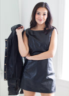 A woman modeling a black faux-leather sleeveless mini dress, holding a matching black faux-leather jacket over her shoulder against a bright, neutral indoor background.