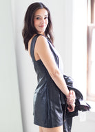 A woman wearing a black faux-leather sleeveless mini dress, holding a matching black jacket in a bright indoor setting.
