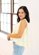 Woman wearing a yellow sleeveless top and blue jeans standing in a bright room with large windows.
