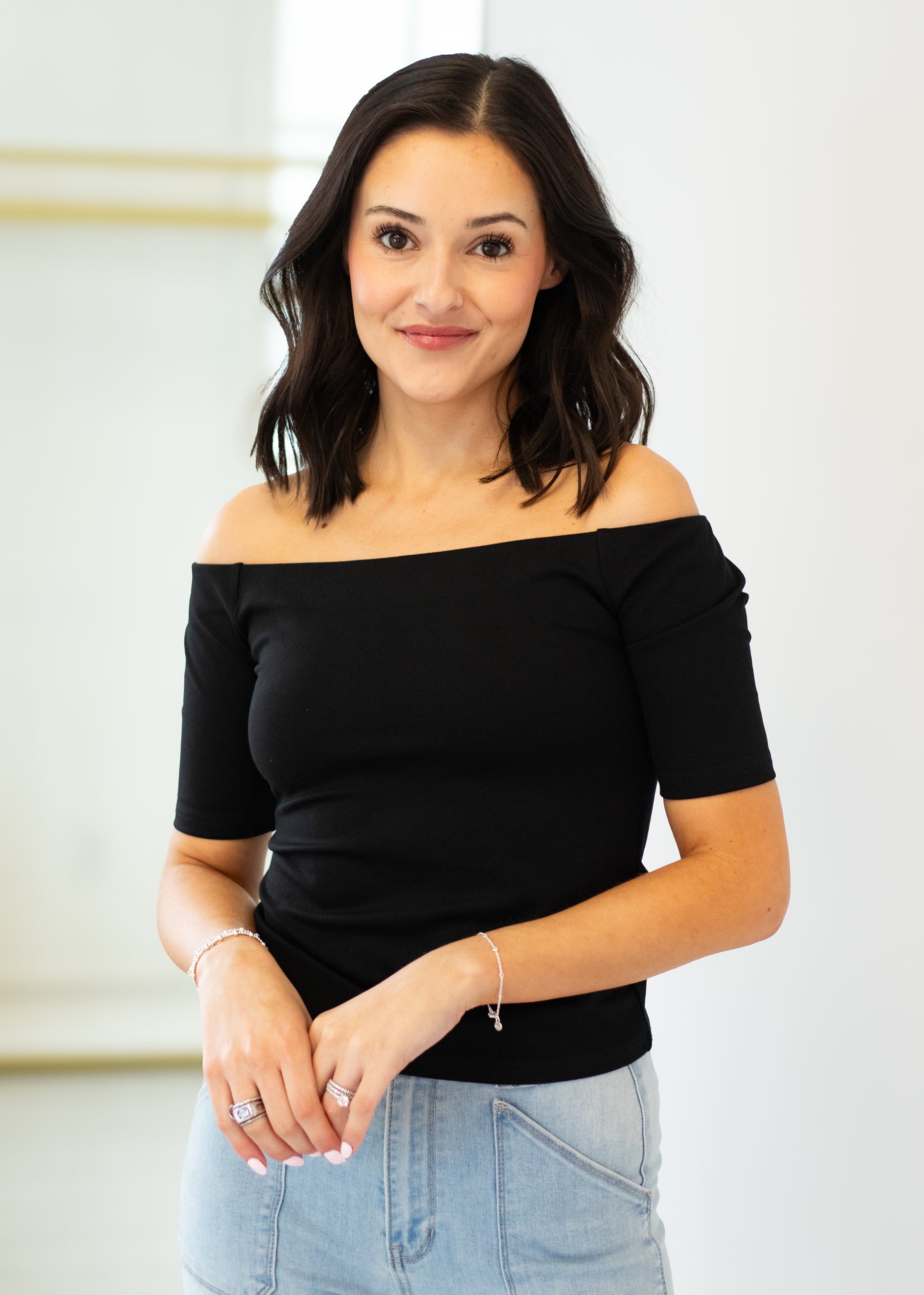 Woman wearing a black off-shoulder top and light blue jeans against a white background