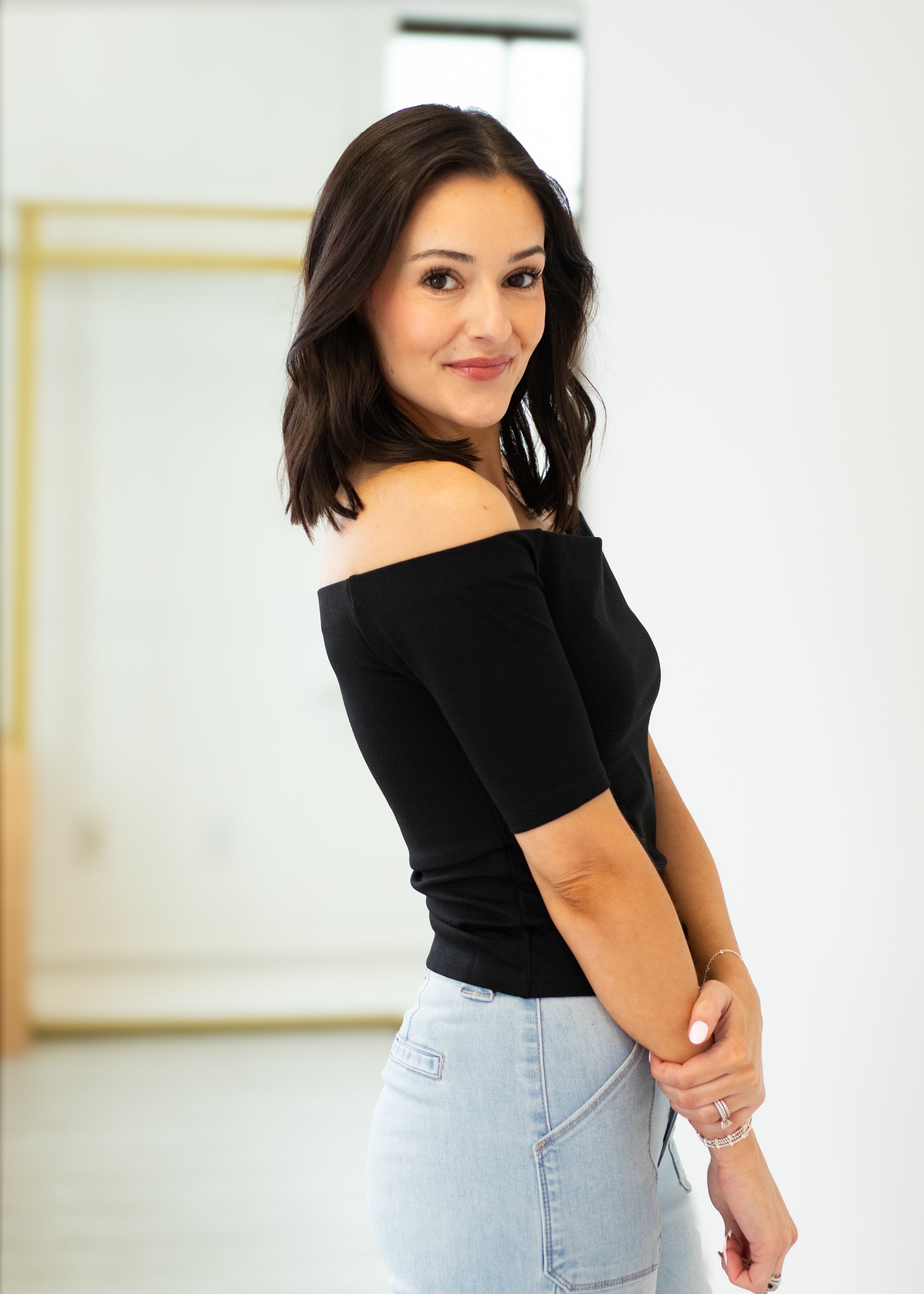 Woman wearing a black off-shoulder top and light blue jeans indoors.