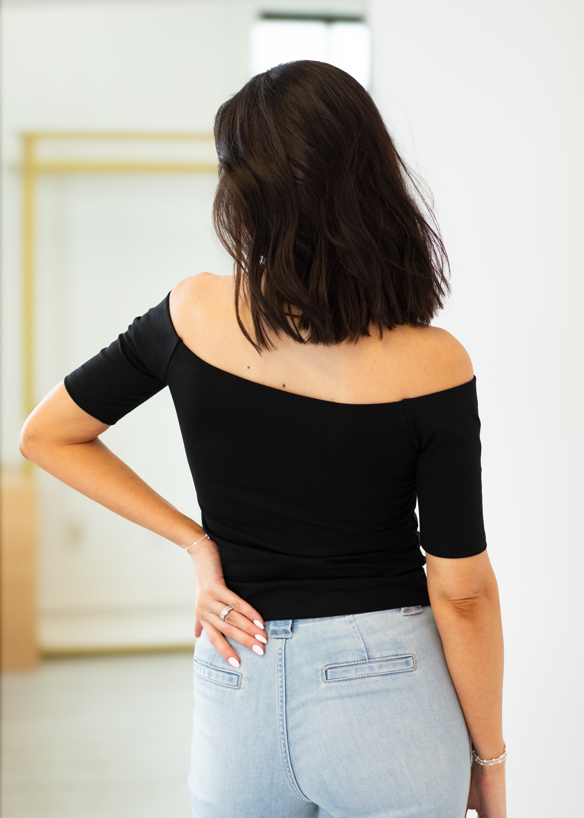 Woman wearing a black off-shoulder top and light blue jeans in an indoor setting.