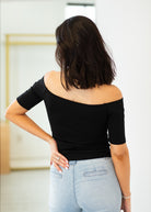 Woman wearing a black off-shoulder top and light blue jeans in an indoor setting.