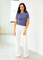 Woman wearing a purple shirt and white pants standing in a minimalistic room.