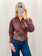 A woman takes a mirror selfie wearing a long-sleeve, button-down brown shirt tucked into blue jeans.