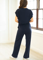 Woman wearing a navy blue outfit standing in a bright room with large windows.