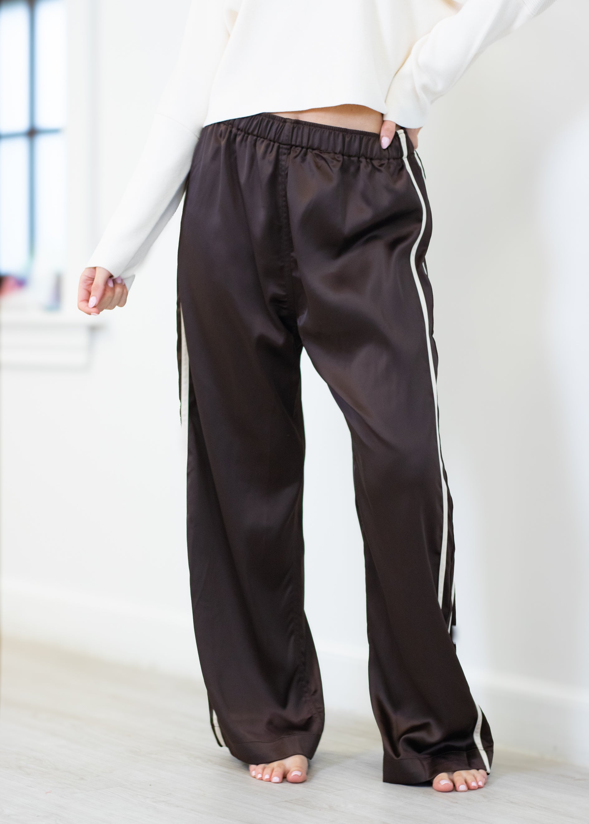 A person wearing dark brown satin-like wide-leg pants with white side stripes, shown from the waist down in a bright indoor room.