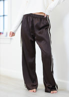 A person wearing dark brown satin-like wide-leg pants with white side stripes, shown from the waist down in a bright indoor room.