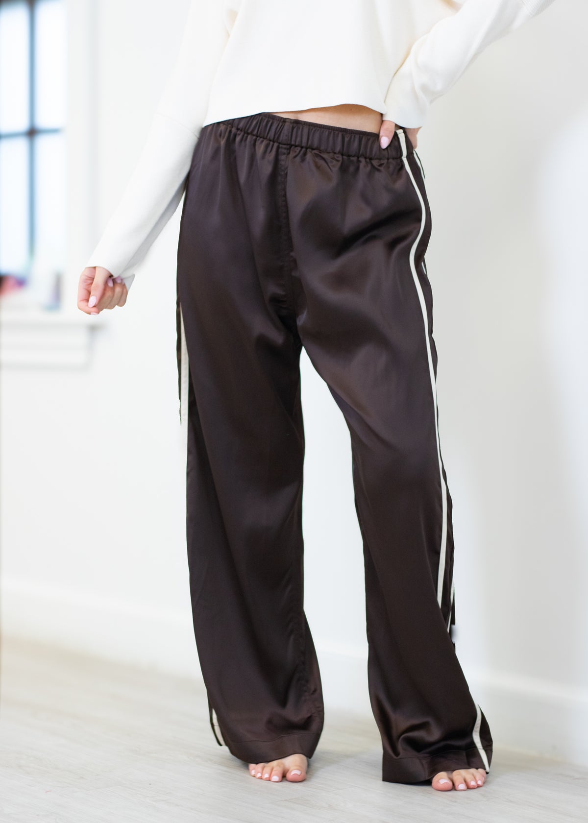 A person wearing dark brown satin-like wide-leg pants with white side stripes, shown from the waist down in a bright indoor room.