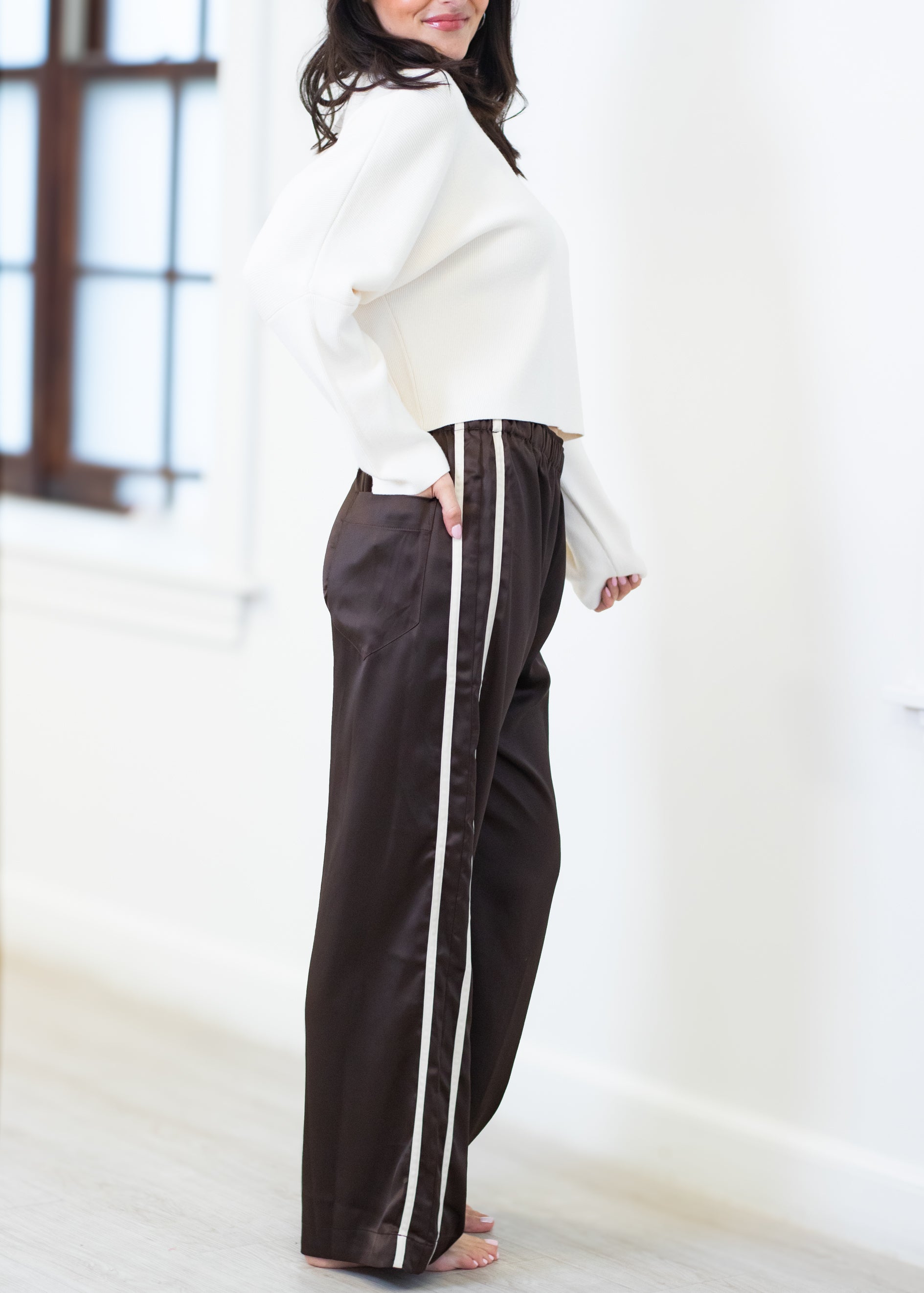 A woman wearing dark brown wide-leg pants with a white side stripe, paired with a white cropped sweater, standing in a bright room.