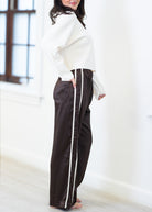 A woman wearing dark brown wide-leg pants with a white side stripe, paired with a white cropped sweater, standing in a bright room.