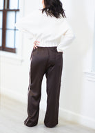 A person shown from the back wearing dark brown wide-leg satin pants with white side stripes and a white knit sweater, standing in a bright indoor room.