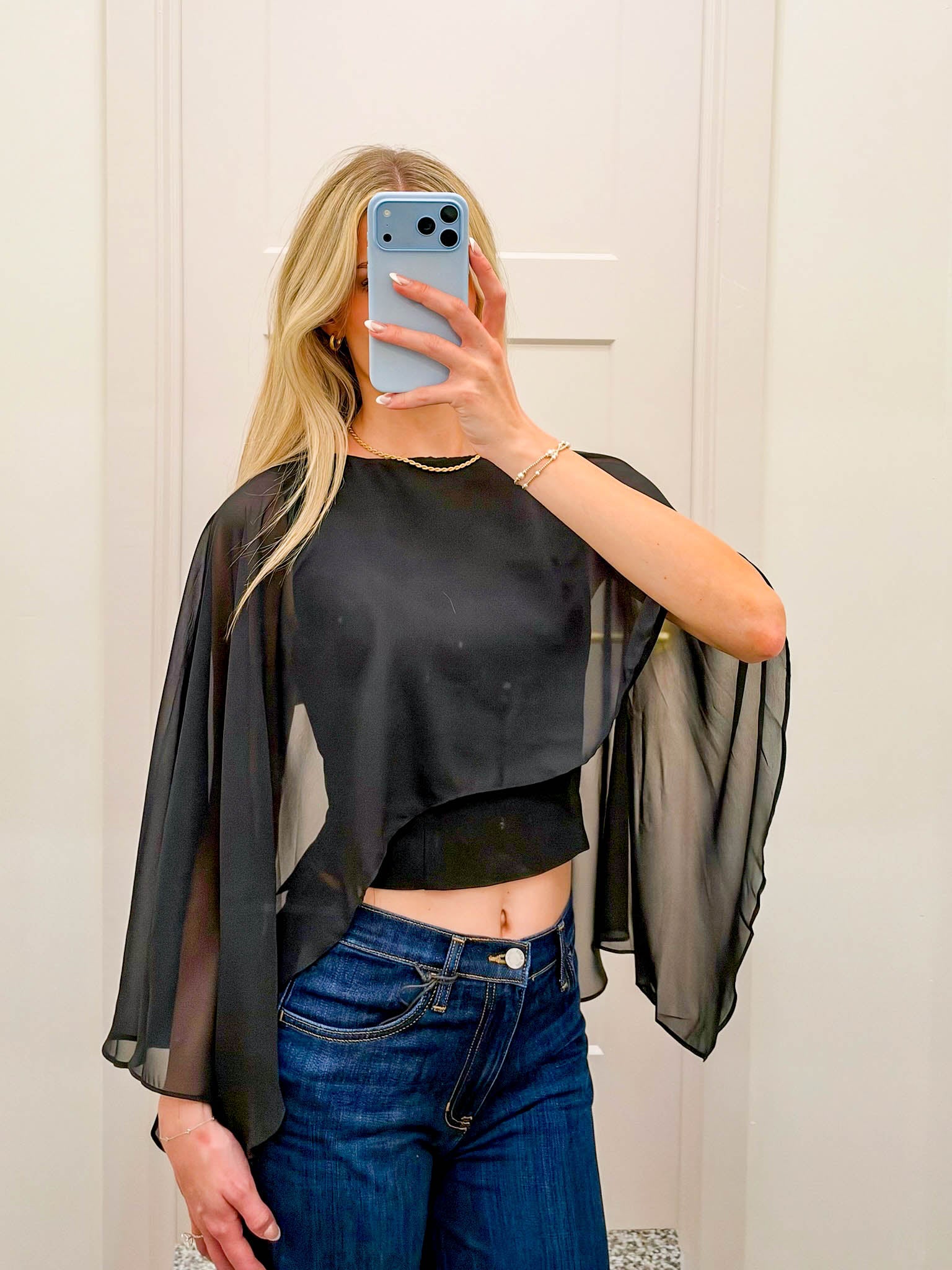 A woman takes a mirror selfie wearing a sheer black long-sleeve crop top with wide draped sleeves and dark blue jeans.