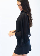 A woman wearing a black sheer cape-sleeve top and matching black shorts, shown from a side profile against a plain white studio background.