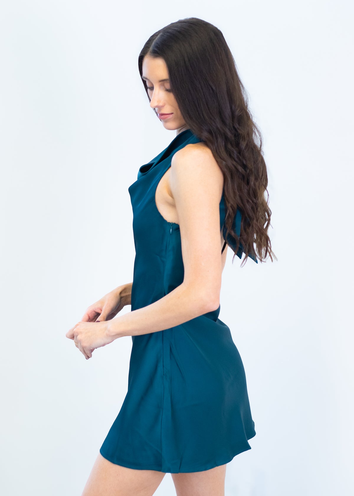 A woman with long dark brown hair is shown from a side-back angle wearing a sleeveless teal blue mini dress against a plain white studio background.