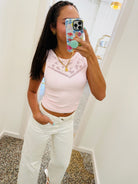 A woman takes a mirror selfie wearing a light pink ribbed tank top with a lace-like V-neckline detail and white pants. She is holding a phone with a floral case and mint green pop socket in a brightly lit indoor room.