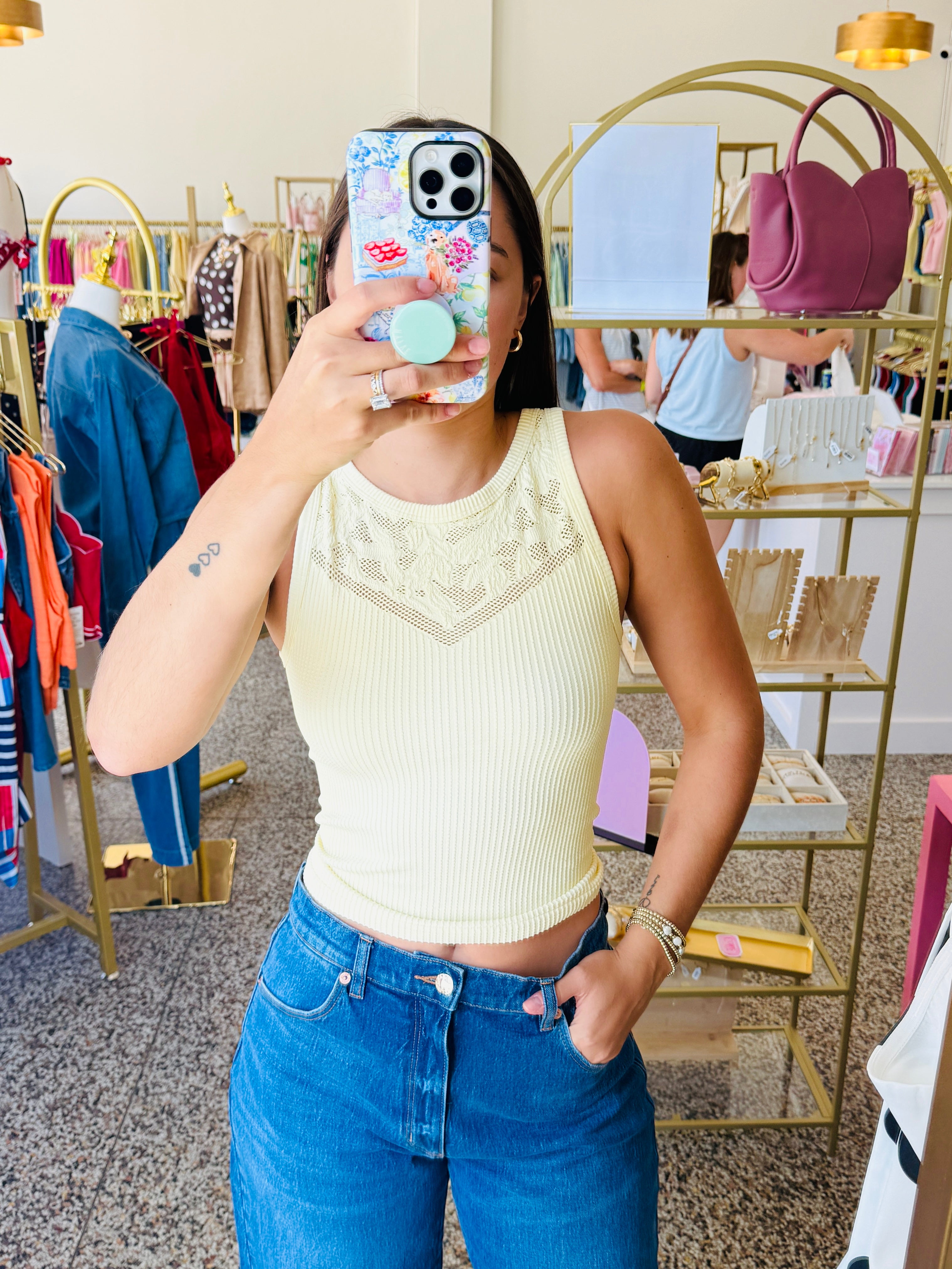 A woman takes a mirror selfie in a boutique, wearing a pale yellow ribbed tank top with a lace V-neckline detail and blue jeans. The boutique has gold clothing racks, a display shelf with a pink handbag, and a speckled floor.