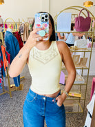A woman takes a mirror selfie in a boutique, wearing a pale yellow ribbed tank top with a lace V-neckline detail and blue jeans. The boutique has gold clothing racks, a display shelf with a pink handbag, and a speckled floor.