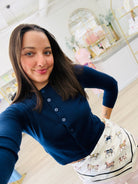 A woman taking a selfie in a boutique, wearing a navy blue long-sleeve button-up cardigan and a white skirt with a horse pattern.