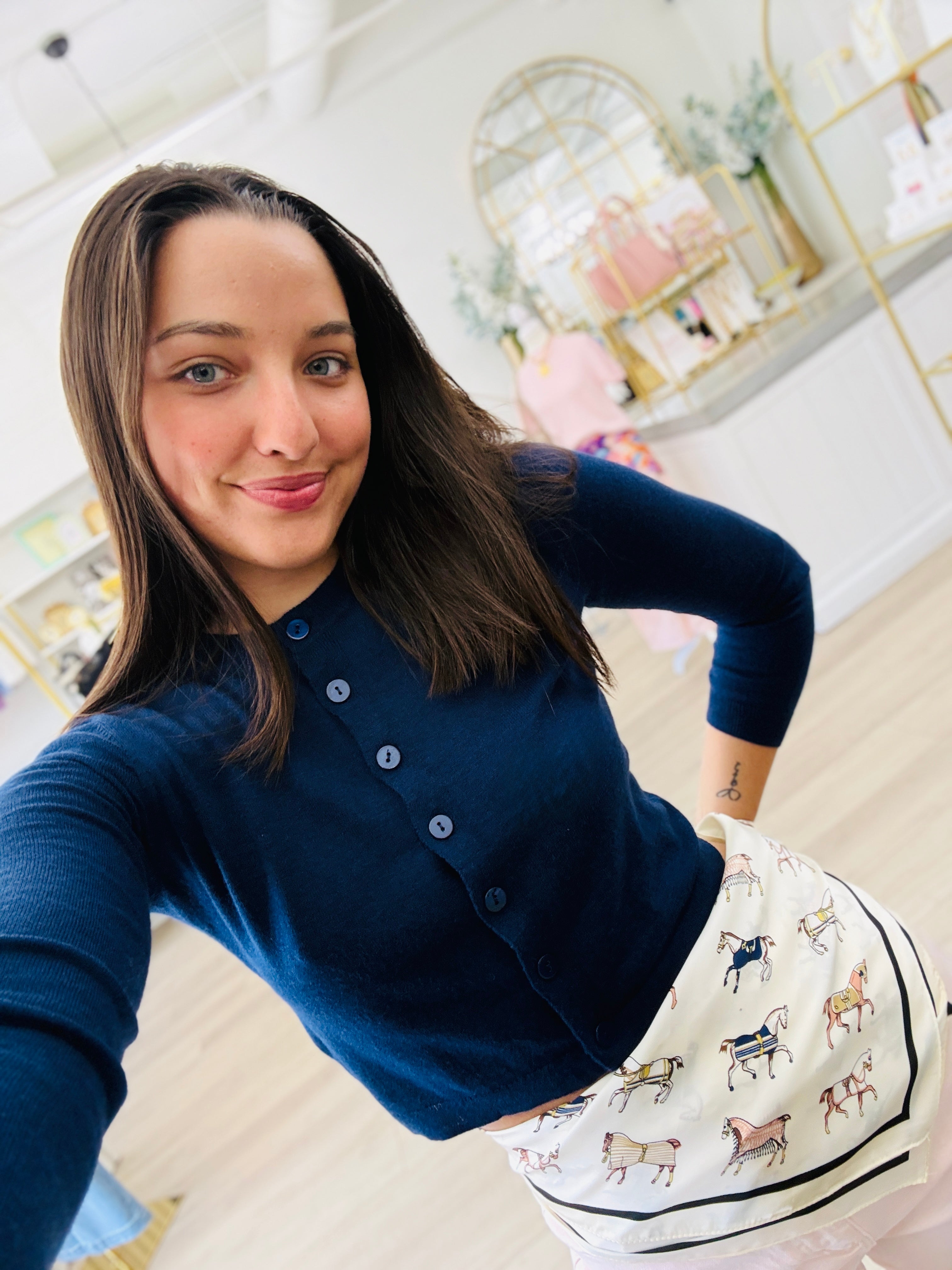 A woman taking a selfie in a boutique, wearing a navy blue long-sleeve button-up cardigan and a white skirt with a horse pattern.