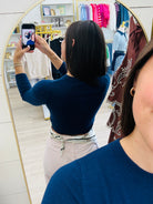A woman takes a mirror selfie in a clothing boutique, wearing a navy blue long-sleeve crop top and light beige pants. The boutique has a gold-framed mirror, light wood floors, and racks of colorful clothing in the background.