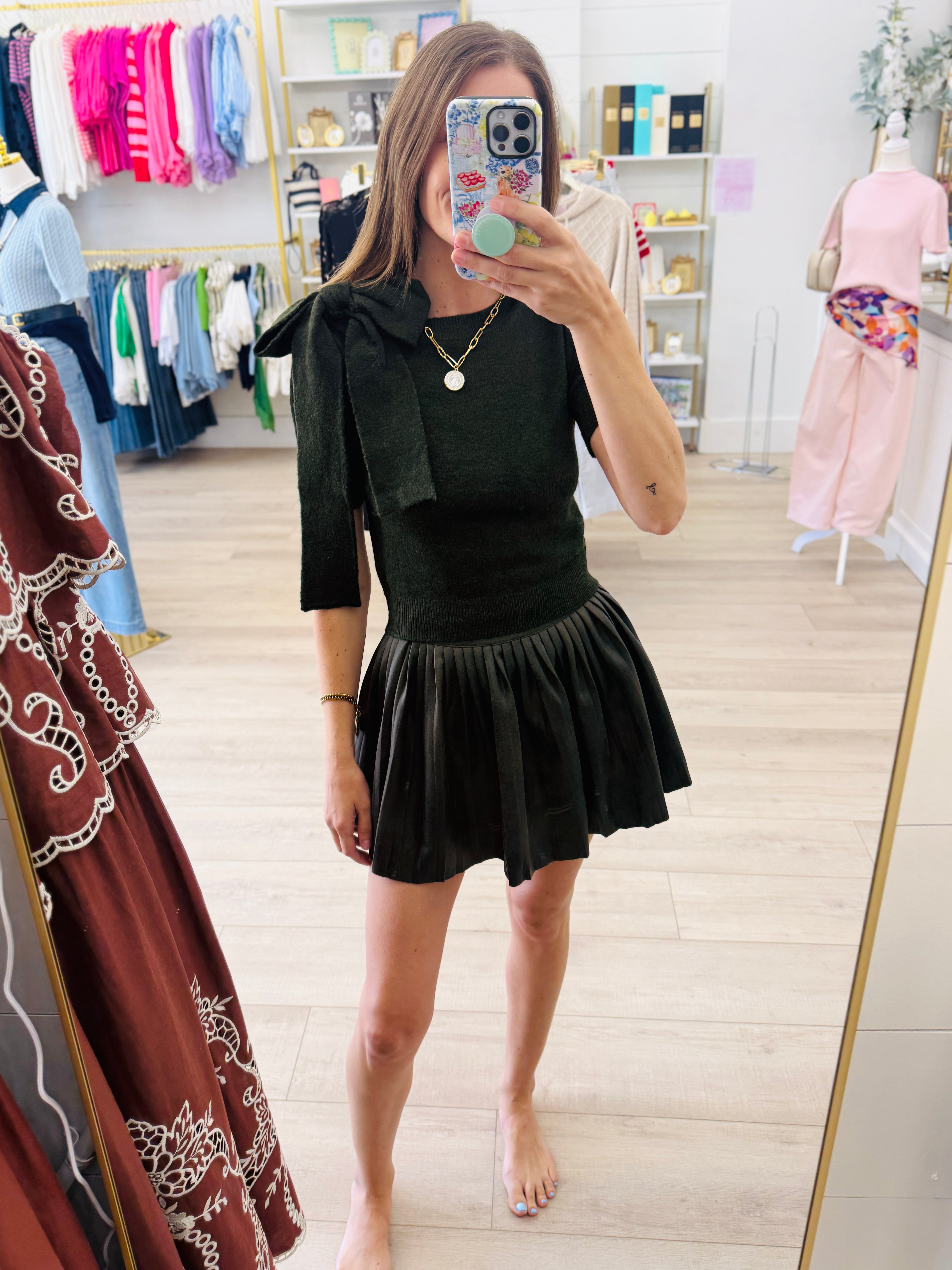 A woman takes a mirror selfie in a boutique while wearing a black velvet short-sleeve top with a large bow detail on the shoulder and a matching black pleated mini skirt.