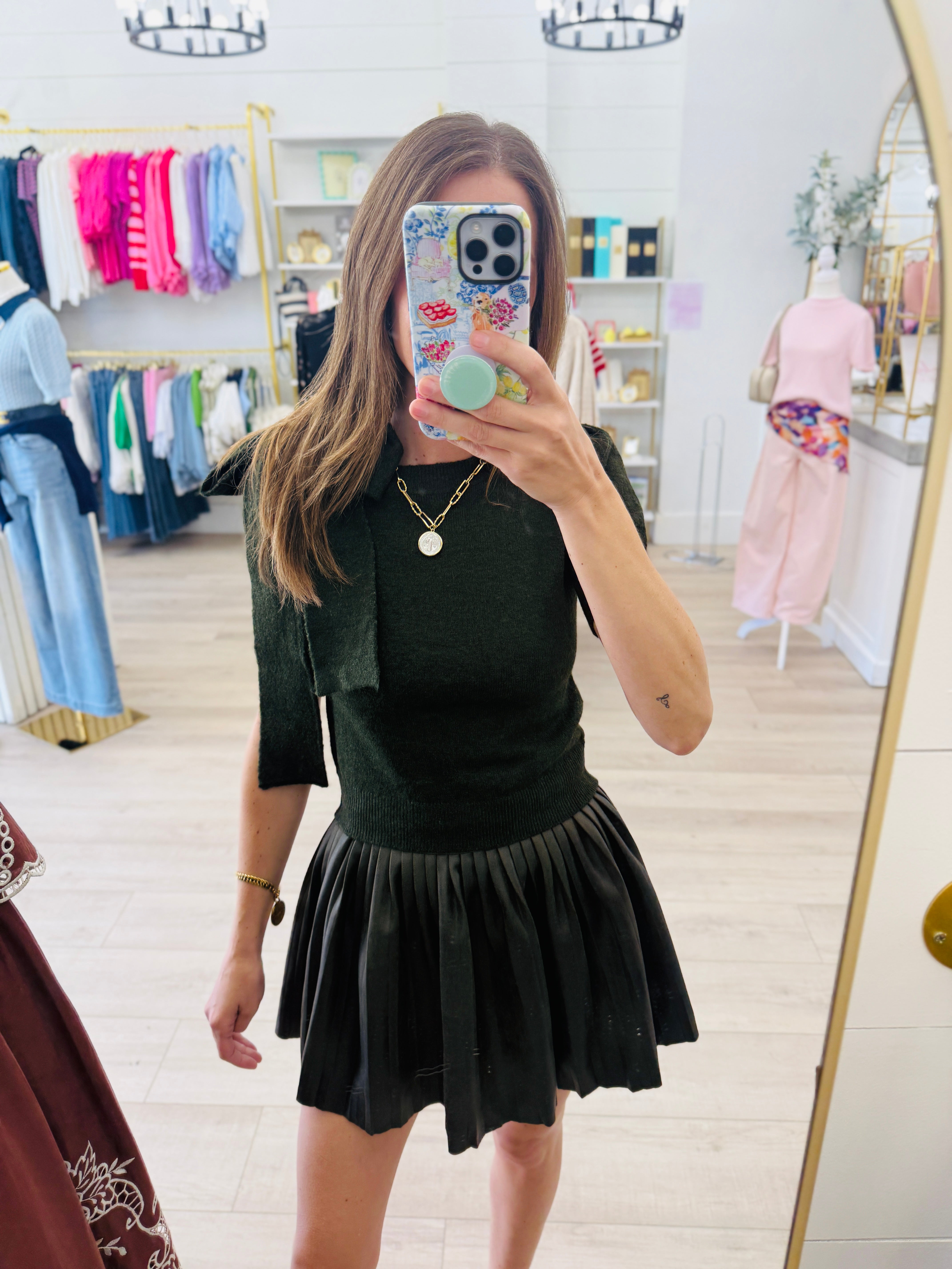 A woman takes a mirror selfie in a clothing boutique, wearing a dark green knit top with a matching pleated black mini skirt.