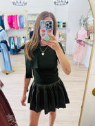 A woman takes a mirror selfie in a clothing boutique, wearing a dark green knit top with a matching pleated black mini skirt.