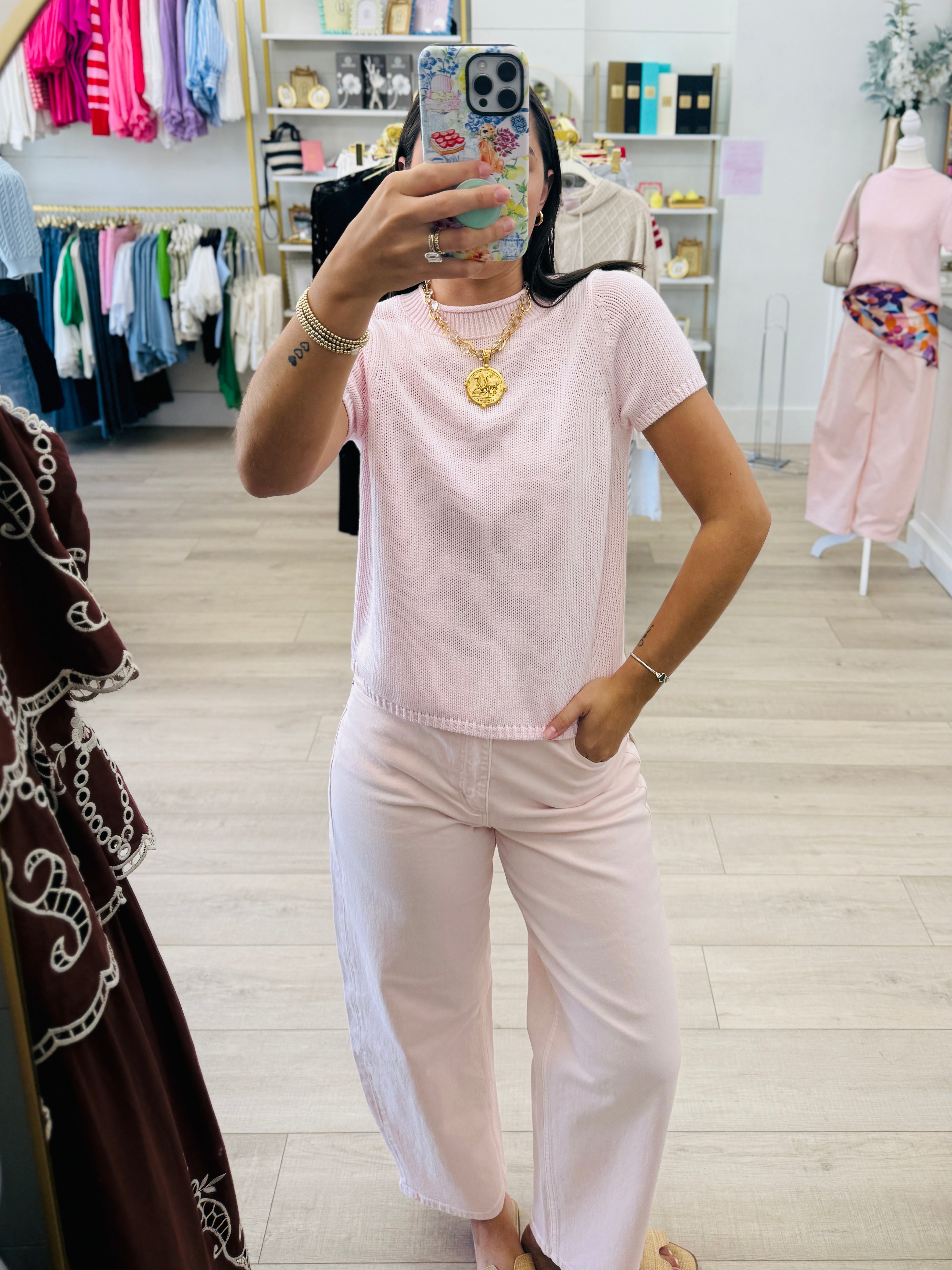 A woman takes a mirror selfie in a boutique while wearing a light pink short-sleeve knit top and matching wide-leg pants. The boutique has light wood floors, clothing racks, and mannequins in the background.
