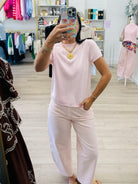 A woman takes a mirror selfie in a boutique while wearing a light pink short-sleeve knit top and matching wide-leg pants. The boutique has light wood floors, clothing racks, and mannequins in the background.