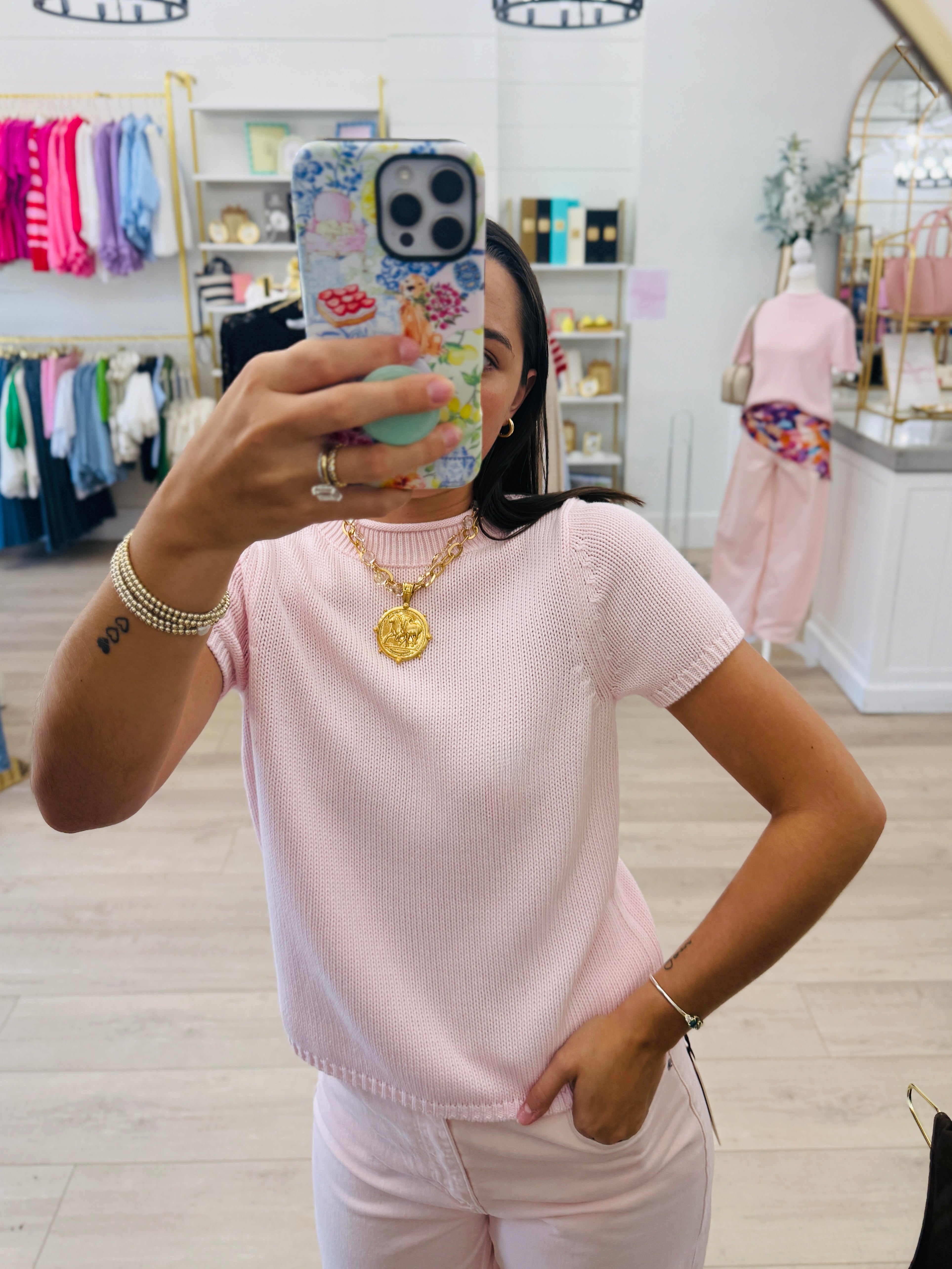 A woman takes a mirror selfie in a boutique while wearing a light pink ribbed short-sleeve top and matching pink pants. She is wearing gold jewelry, including a gold medallion necklace, and holding a floral-patterned phone case.