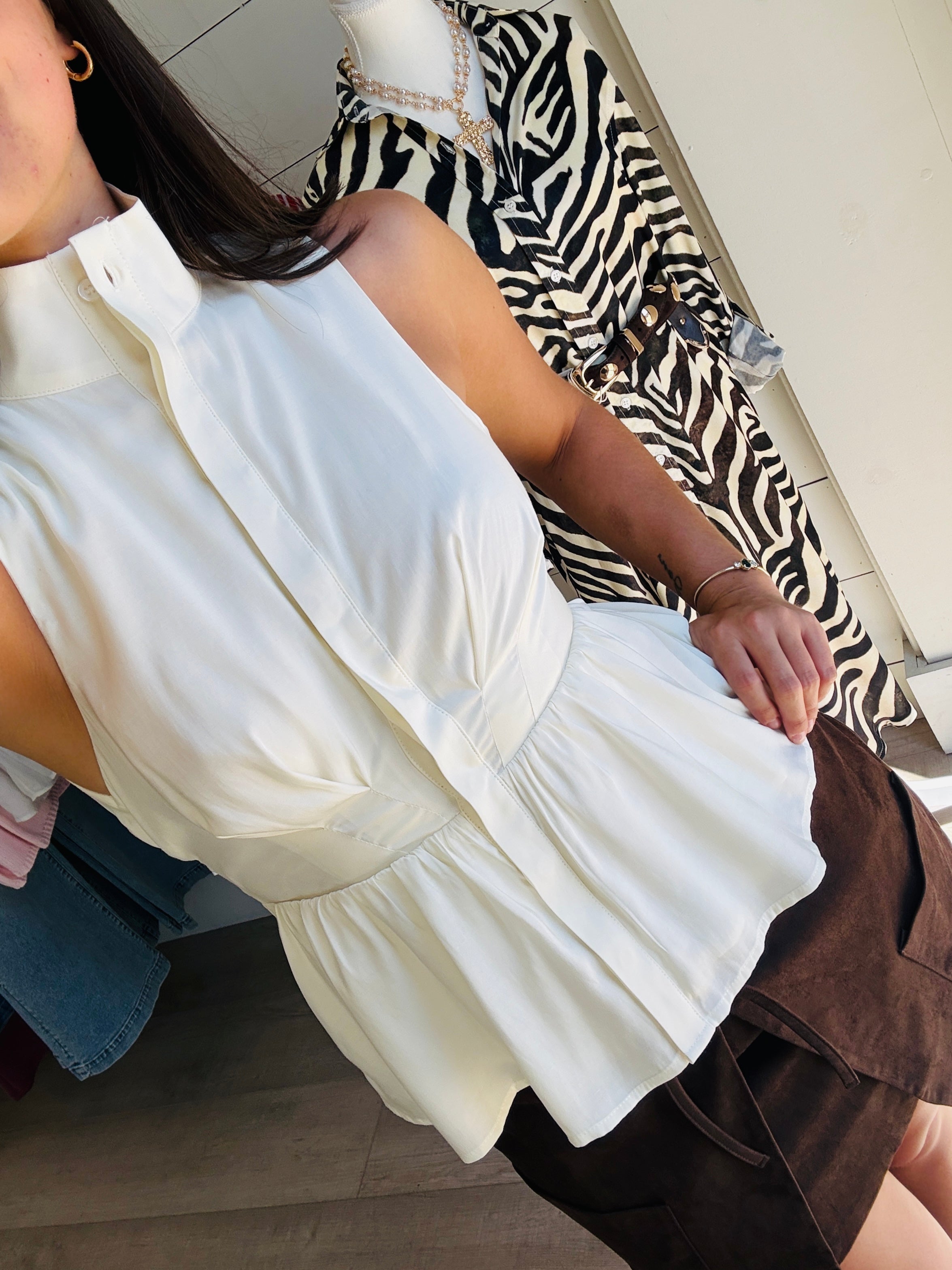 A woman wearing a sleeveless white peplum blouse with a high collar and gathered waist, paired with a brown skirt. The photo is taken in a mirror within a clothing store, showing other garments in the background.