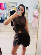 A woman takes a mirror selfie in a boutique wearing a dark brown sleeveless button-front top and matching skirt set. The boutique has light wood floors, clothing racks, and mannequins in the background.