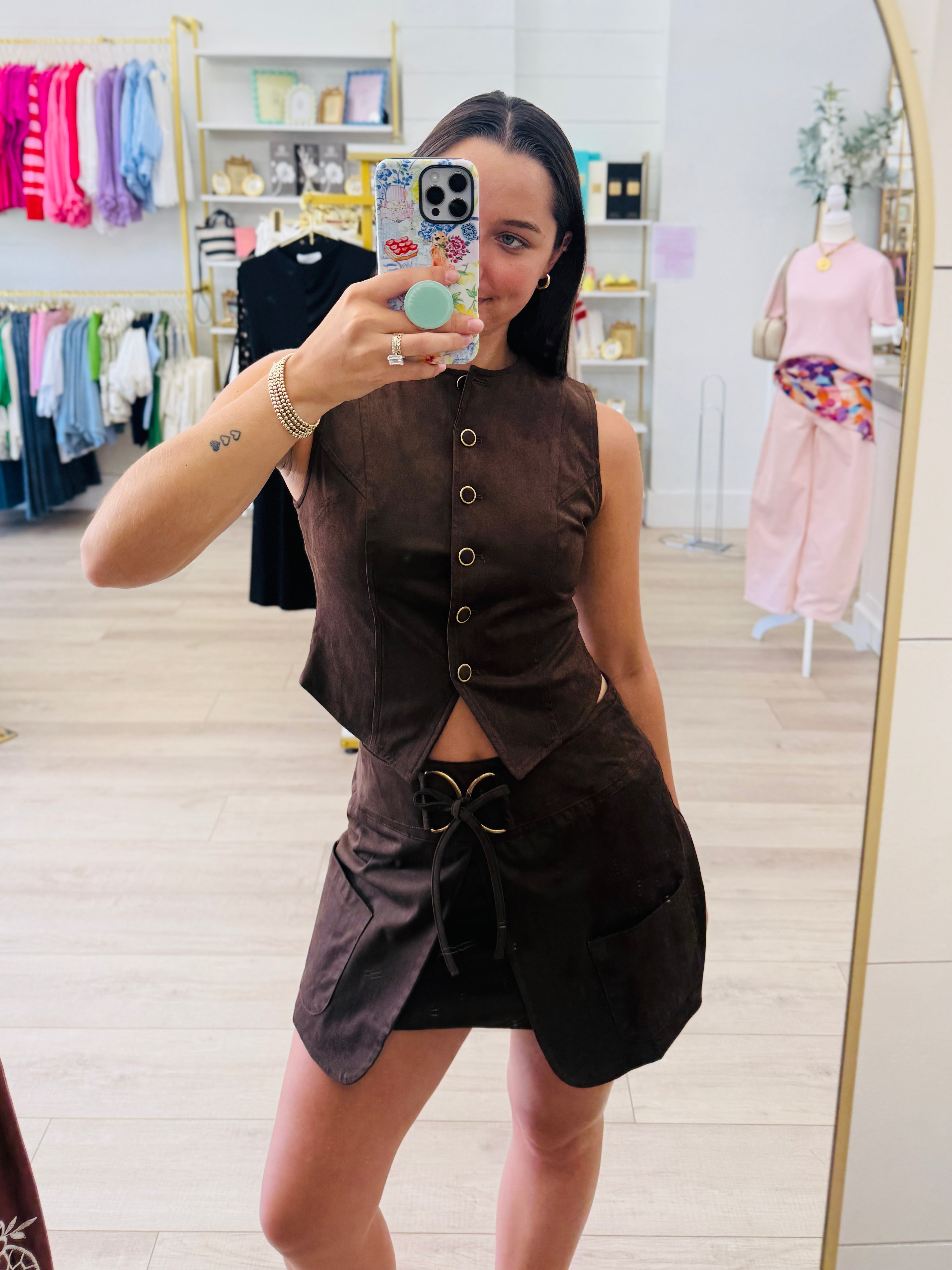 A woman takes a mirror selfie in a boutique wearing a dark brown sleeveless button-front top and matching skirt set. The boutique has light wood floors, clothing racks, and mannequins in the background.