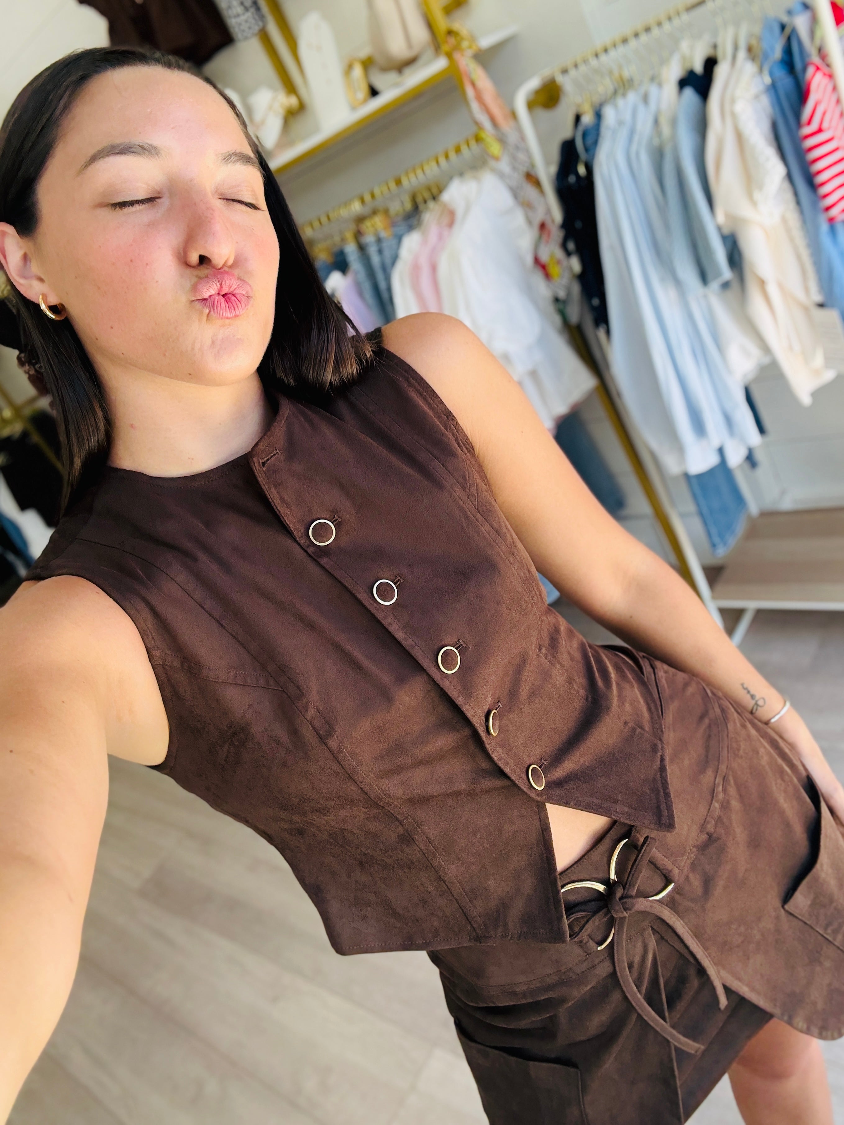 A woman takes a selfie wearing a brown sleeveless vest and matching skirt set in a boutique clothing store. The outfit features a buttoned front on the vest and a tie detail on the skirt.