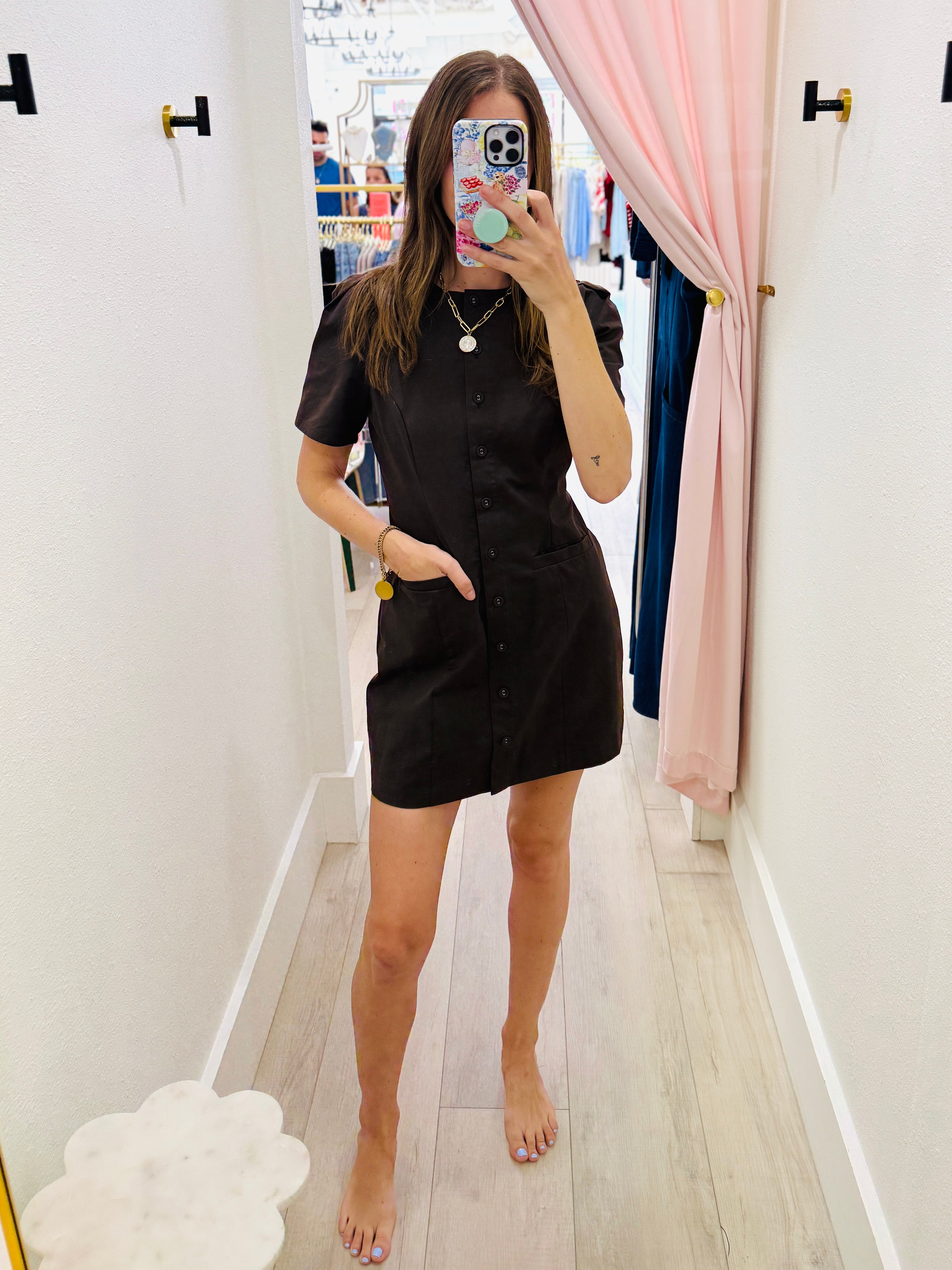 A woman takes a mirror selfie in a boutique wearing a black short-sleeve button-front shirt dress. The boutique has white walls, light wood floors, and pink curtains.