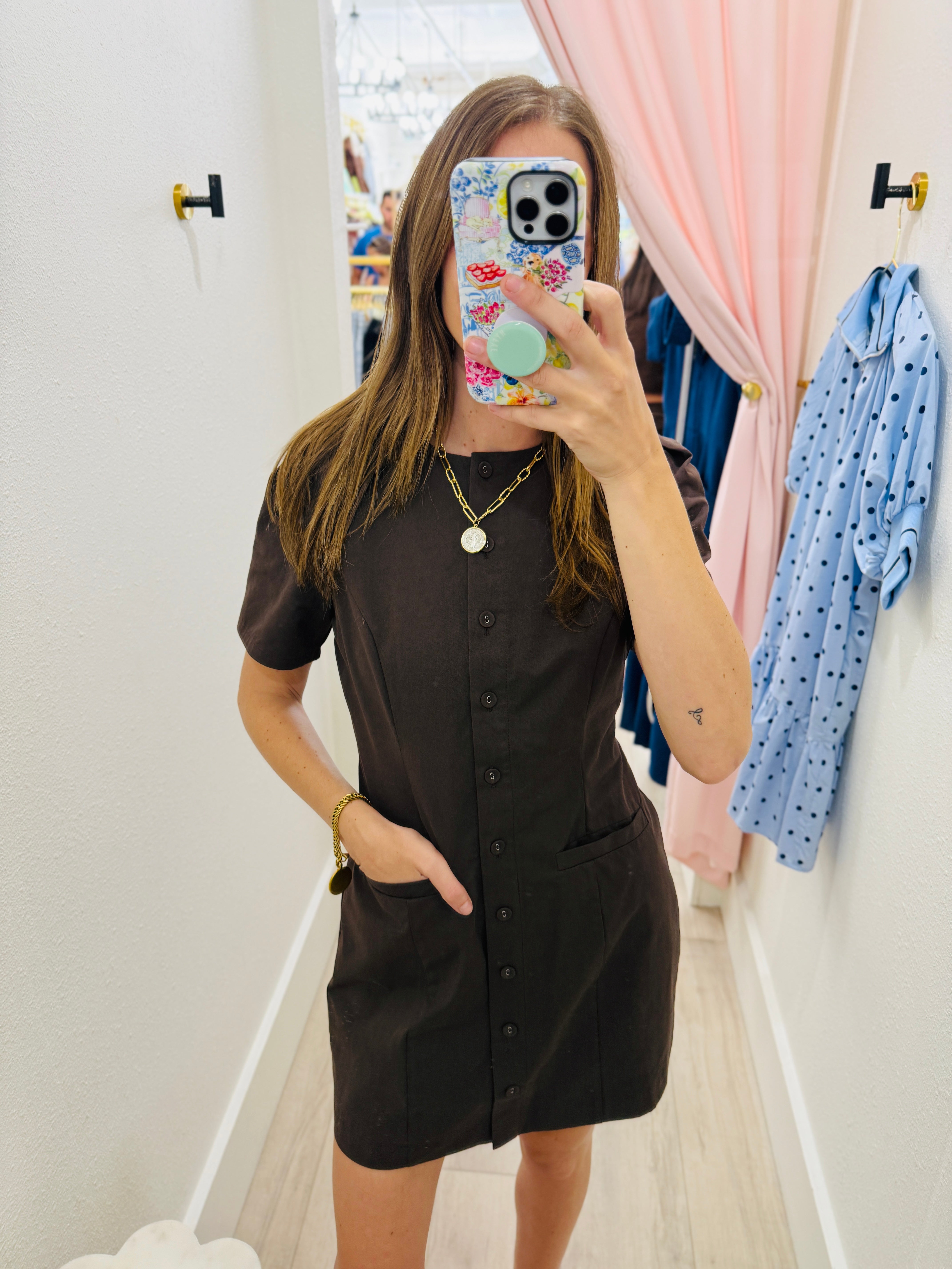 A woman takes a mirror selfie in a fitting room wearing a dark brown short-sleeve button-front dress with pockets. A pink curtain and a blue polka-dot shirt are visible in the background.