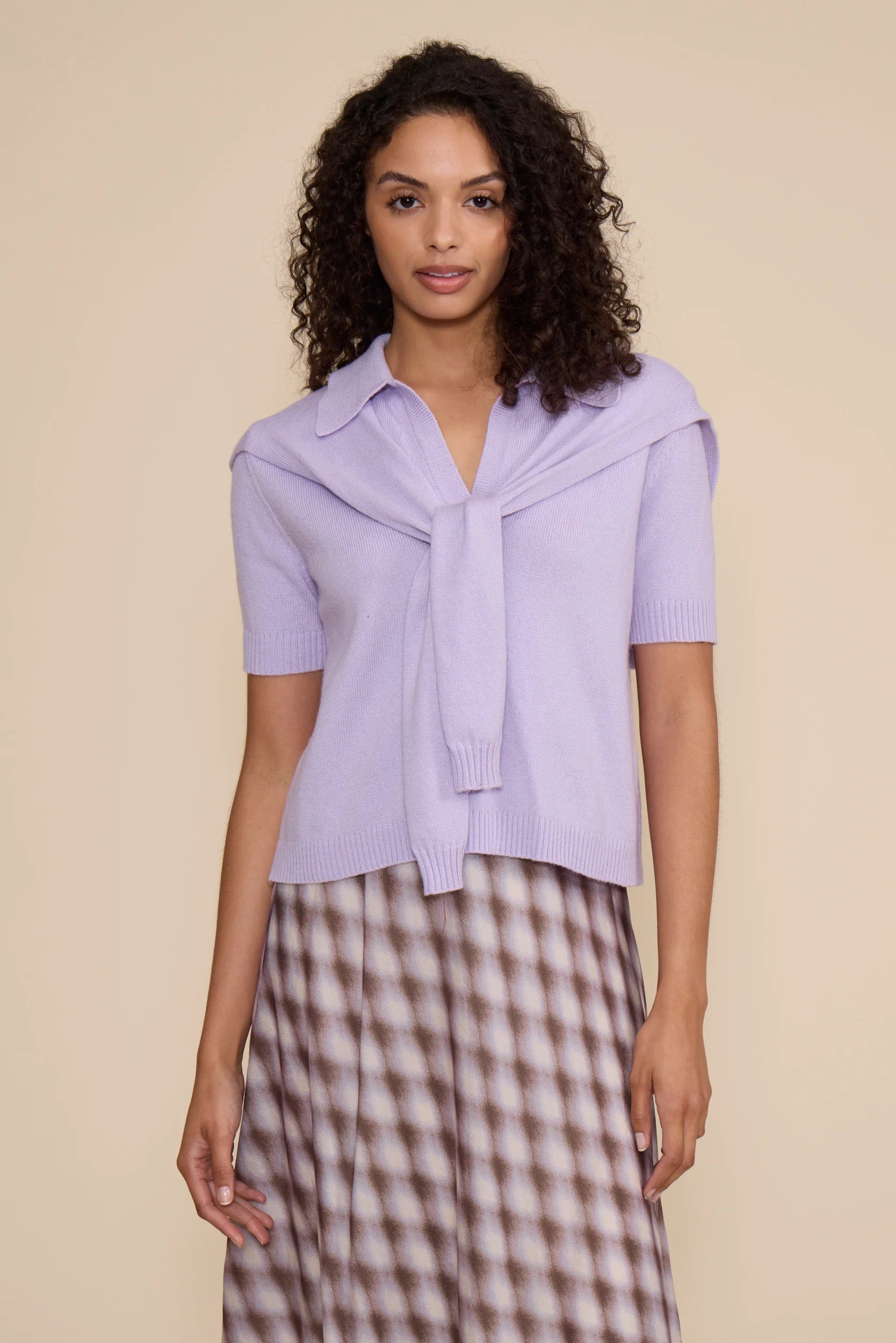 A model wearing a lavender short-sleeve knit top with a large collar and front tie detail, paired with a brown and white patterned skirt, against a solid beige background.