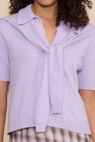 A close-up of a person wearing a light lavender knit short-sleeve top with a wide collar and a front tie detail, paired with a brown-and-white patterned skirt against a plain beige background.