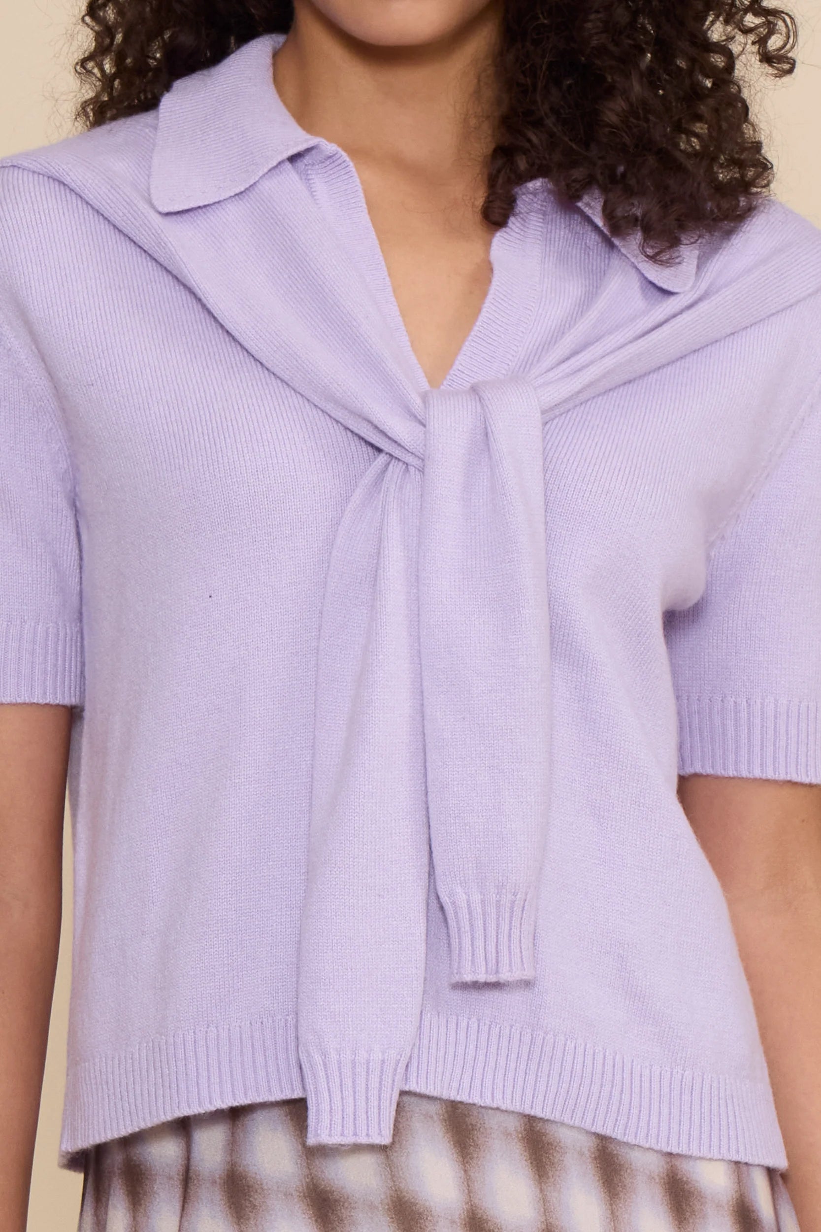 A close-up of a person wearing a light lavender knit short-sleeve top with a wide collar and a front tie detail, paired with a brown-and-white patterned skirt against a plain beige background.