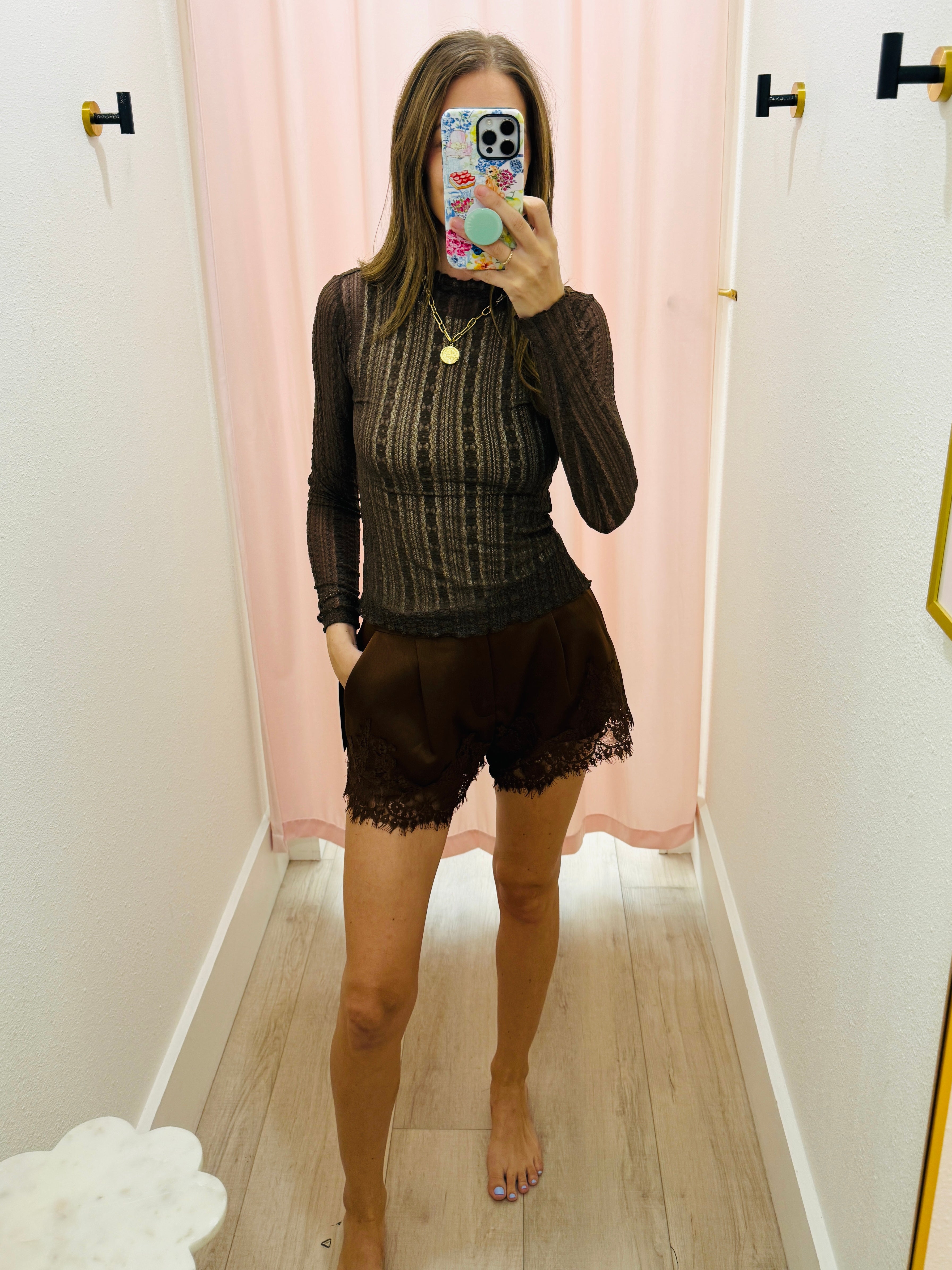 A woman takes a mirror selfie in a fitting room wearing a dark brown ribbed long-sleeve top and matching dark brown lace-trimmed shorts.