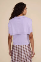 A woman shown from the back wearing a light lavender knit cropped shrug or cape with short sleeves, paired with a brown and white patterned skirt, against a solid beige background.