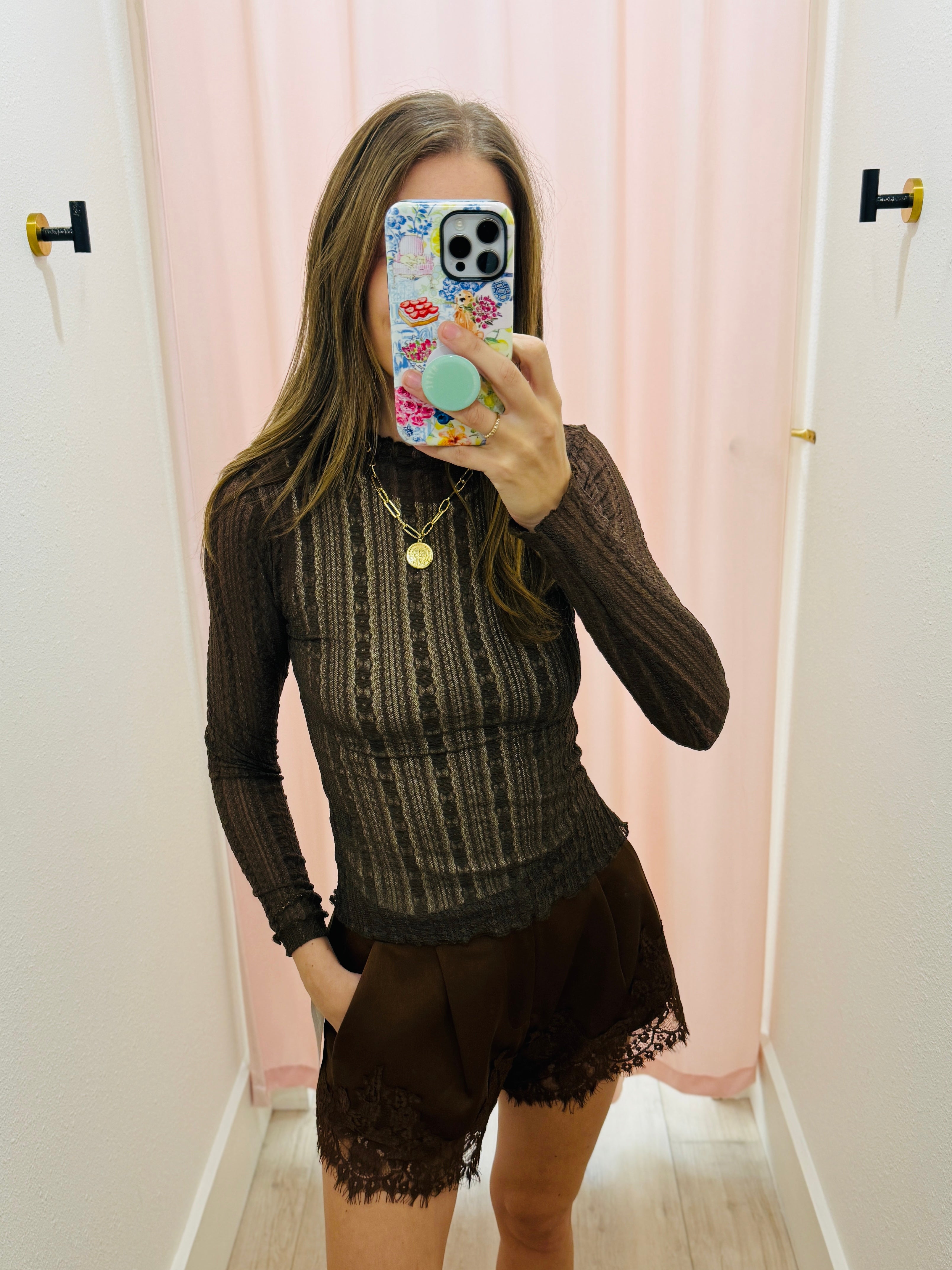 A woman takes a mirror selfie in a fitting room wearing a dark brown ribbed long-sleeve knit top and matching brown lace-trimmed shorts. She is holding a phone with a floral case, with a pink curtain and white walls in the background.