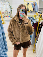 A woman takes a mirror selfie in a clothing boutique wearing a brown fuzzy faux fur jacket and brown lace-up shorts.