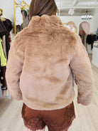 A person viewed from behind wearing a plush faux-fur cropped jacket in a warm taupe-brown shade, paired with brown lace-trim shorts inside a boutique setting.