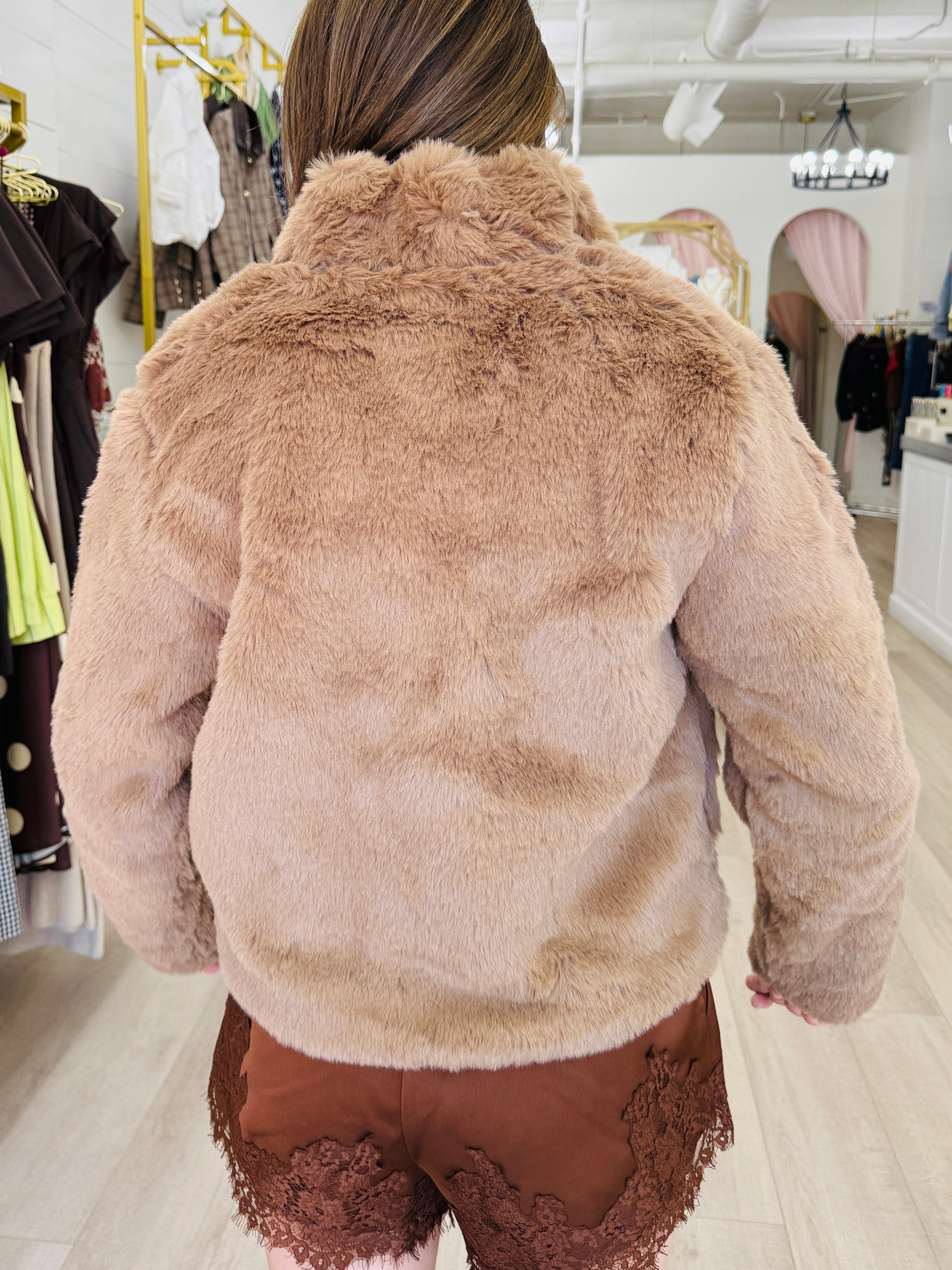 A person viewed from behind wearing a plush faux-fur cropped jacket in a warm taupe-brown shade, paired with brown lace-trim shorts inside a boutique setting.