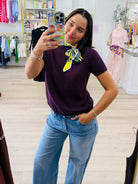 A woman takes a mirror selfie in a boutique, wearing a short-sleeve purple knit sweater and light blue jeans, with a colorful patterned neck scarf tied around her collar.