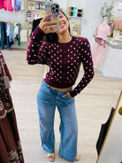 A woman takes a mirror selfie in a clothing boutique, wearing a dark burgundy long-sleeve cardigan with white polka dots and light-wash blue wide-leg jeans.